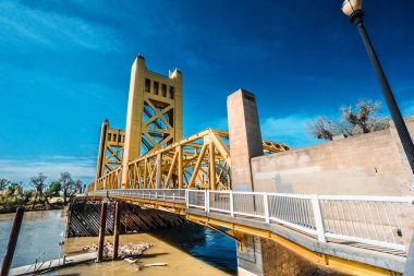 Sarı renkli ve beyaz korkuluklu bir köprü. Köprü bir nehrin üzerinde ve su kahverengi.