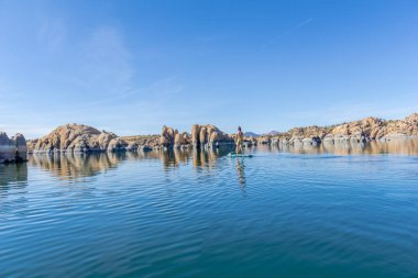 Bir adam kayalarla çevrili bir gölde kürek çekiyor. Su sakin ve mavidir, ve gökyüzü berrak ve parlaktır. Huzur ve macera kavramı