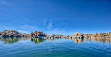 Kayalık bir kıyı şeridi olan güzel mavi bir göl. Su sakin ve durgun, yukarıdaki gökyüzünü yansıtıyor. Sahne huzurlu ve sakin, kayalar ve su bir huzur hissi yaratıyor.