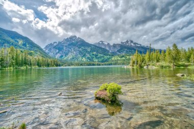 Arkasında dağlar olan bir göl. Su sakin ve berrak. Gökyüzü bulutlu, ama dağlar uzaklardan görülebilir.