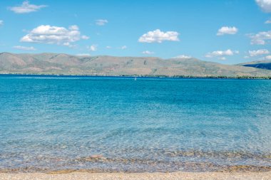 Gökyüzü açık, güzel mavi bir okyanus. Su sakin ve huzurlu. Arka plandaki dağlar dingin atmosfere katkı sağlar.
