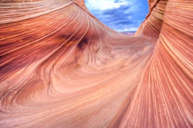 Derin, kıvrımlı bir duvarı olan büyük bir kanyon. Kanyon kırmızı, turuncu ve kahverengi olmak üzere çeşitli renklerle doludur. Kanyonun üstündeki gökyüzü bulutlu, derinlik hissi katıyor.