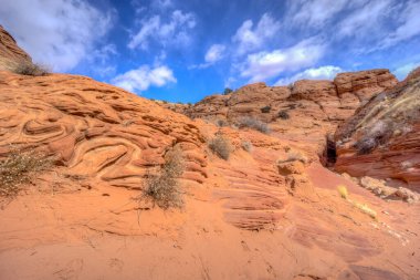 Kayalık yamaçlı bir çöl manzarası ve bulutlu bir gökyüzü. Gökyüzü mavi ve güneş parlıyor.