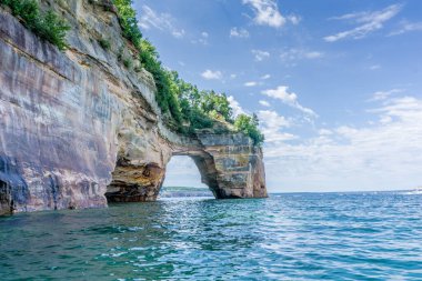 Arka planda büyük bir kaya oluşumu olan güzel mavi bir okyanus. Kaya oluşumu ağaçlarla çevrili büyük bir kemerdir. Su sakin ve gökyüzü berrak.