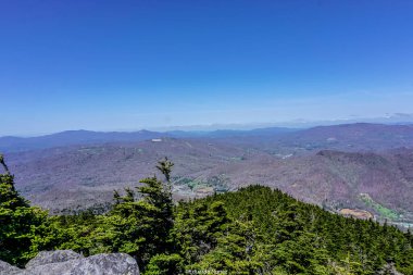 Arka planda açık mavi gökyüzü olan bir dağ sırası. Dağlar ağaçlarla kaplı ve manzara nefes kesici.