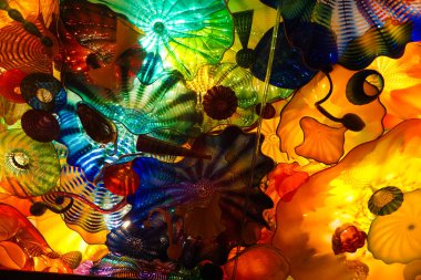 Sarı merkezli renkli cam çiçekler sergiliyor. Çiçekler bir hareket ve derinlik hissi yaratacak şekilde düzenlenmiştir. Renkler canlı ve göz alıcı.