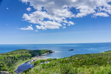 Ortasında küçük bir ada olan güzel bir göl manzarası. Gökyüzü açık ve mavidir. Birkaç bulut etrafa saçılmıştır. Su sakin ve gölün etrafındaki ağaçlar yemyeşil ve yemyeşil.