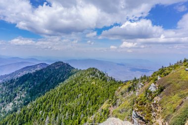 Arka planda bulutlu bir gökyüzü olan bir dağ sırası. Bulutlar beyaz ve kabarık, ve gökyüzü mavi. Dağlar ağaçlarla kaplı ve manzara nefes kesici.