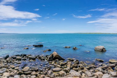Arka planda sakin mavi bir okyanus olan kayalık bir kıyı şeridi. Kayalar kıyı şeridine dağılmış, resim gibi bir manzara yaratıyor.