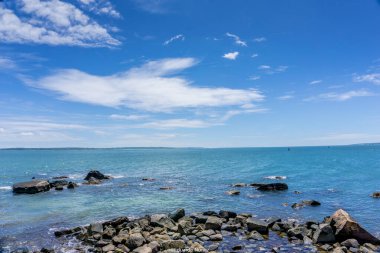Okyanus sakin ve gökyüzü açık. Kıyıdaki kayalar dağılmış ve su mavi.