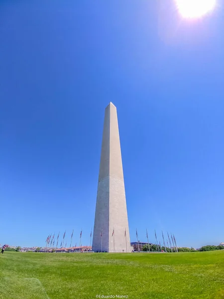 Washington Anıtı çimenli bir arazinin ortasında uzun beyaz bir yapıdır. Gökyüzü açık ve mavidir, ve güneş parlıyor. Anıt bayraklarla çevrili.