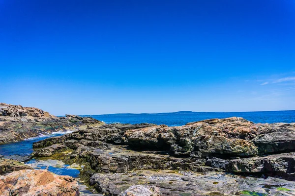 Arka planda mavi gökyüzü olan kayalık bir kıyı şeridi. Kayalar dağılmış ve su sakin.