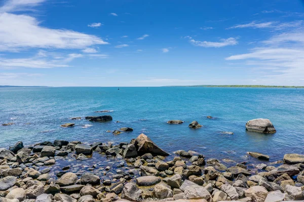 Arka planda sakin mavi bir okyanus olan kayalık bir kıyı şeridi. Kayalar kıyı şeridine dağılmış, resim gibi bir manzara yaratıyor.