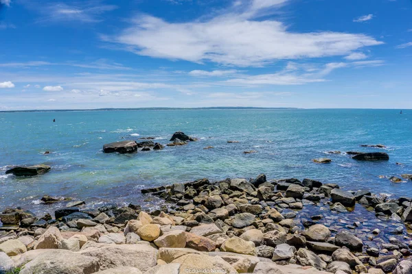 Arka planda sakin bir okyanus olan kayalık bir kıyı şeridi. Kayalar kıyı şeridi boyunca dağılmış, resim gibi bir manzara yaratıyor. Gökyüzü açık ve mavi, birkaç bulut yüzüyor.