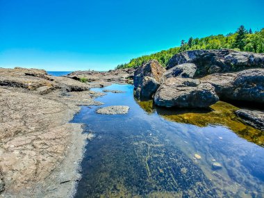 Küçük bir su kütlesi olan kayalık bir kıyı şeridi. Su berrak ve sakin, yukarıdaki mavi gökyüzünü yansıtıyor. Sahne huzurlu ve sakin, kayalar su arasında doğal bir bariyer oluşturuyor.