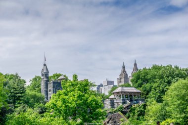 Arkasında büyük bir kale olan bir şehir manzarası. Gökyüzü bulutlu ve ağaçlar yeşil