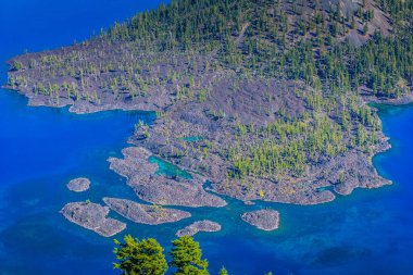Arka planda su kütlesi olan mavi ve yeşil bir ada. Ada ağaçlarla çevrili ve kayalık bir kıyı şeridi var.