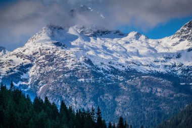 Dağlar karla kaplı ve gökyüzü bulutlu. Huzurlu ve huzurlu bir manzara, ağaçların tepesinde yükselen dağlarla.