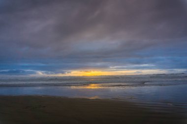 Gökyüzü bulutlu ve güneş batıyor. Okyanus sakin ve su gökyüzünü yansıtıyor.