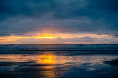 Okyanus sakin ve gökyüzü derin bir mavi. Güneş batıyor ve gökyüzü bulutlarla dolu.