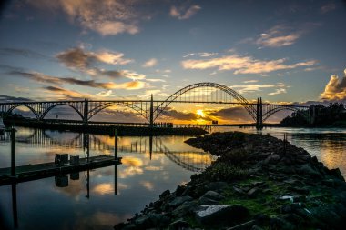 Köprü, arka planda günbatımı olan bir nehri kapsar. Köprü güneş tarafından aydınlatılır ve sıcak ve huzurlu bir atmosfer yaratır.