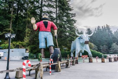 Sakallı ve kırmızı gömlekli büyük bir adam heykeli büyük bir boğa heykelinin yanında duruyor. Sahne, arka planda ağaçların olduğu bir parkta ya da halka açık bir alanda geçiyor.