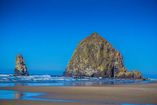 Büyük bir kaya sahilde daha küçük bir kayanın yanında oturur. Gökyüzü berrak ve mavi, ve su sakin. Sahne huzurlu ve sakin, kayalar istikrar ve güç hissi veriyor.