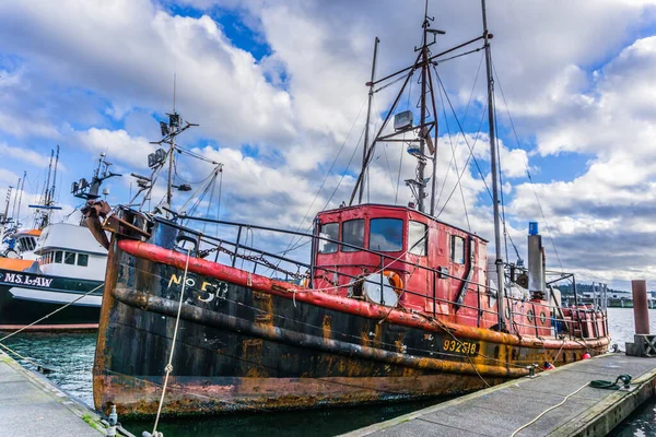 Üzerinde 5 numara olan kırmızı bir tekne marinada duruyor. Tekne eski ve paslı ve diğer teknelerle çevrili. Sahnenin nostaljik ve melankolik bir havası var.