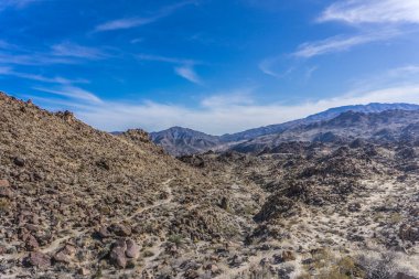 Açık mavi gökyüzü olan kayalık bir çöl manzarası. Gökyüzü bulutlarla kaplı ve güneş ışıl ışıl parlıyor. Sahne, kayaların engin genişliğiyle huzurlu ve huzurlu.