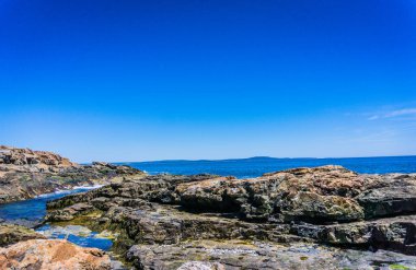 Arka planda mavi gökyüzü olan kayalık bir kıyı şeridi. Kayalar dağılmış ve su sakin.