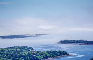 Arka planda ağaçlar ve göl olan sisli bir gün. Gökyüzü birkaç bulutla daha açık.