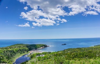 Ortasında küçük bir ada olan güzel bir göl manzarası. Gökyüzü açık ve mavidir. Birkaç bulut etrafa saçılmıştır. Su sakin ve gölün etrafındaki ağaçlar yemyeşil ve yemyeşil.