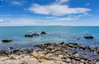 Arka planda sakin bir okyanus olan kayalık bir kıyı şeridi. Kayalar kıyı şeridi boyunca dağılmış, resim gibi bir manzara yaratıyor. Gökyüzü açık ve mavi, birkaç bulut yüzüyor.