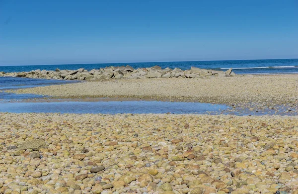 Arka planda su bulunan kayalık bir sahil. Kumsal kayalıktır ve etrafa dağılmış bir sürü kaya vardır. Su sakin ve mavi.