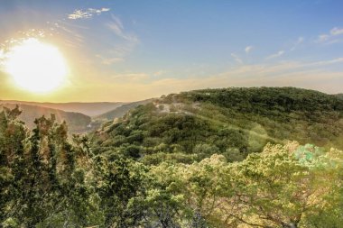 Gökyüzünde parlak bir güneş olan güzel bir dağ manzarası. Güneş batıyor, ağaçların ve tepelerin üzerine sıcak bir parıltı saçıyor. Huzurlu ve huzurlu bir sahne.