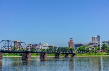 Üstü açık mavi gökyüzü olan bir şehirde bir nehir boyunca uzanan bir köprü. Şehir hareketlilikle dolup taşıyor ve su sakin.
