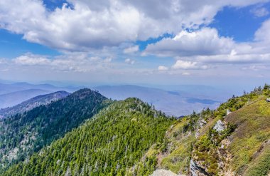 Açık mavi gökyüzü ve birkaç bulutu olan bir dağ sırası. Dağlar ağaçlarla kaplı ve gökyüzü aydınlık ve güneşli.