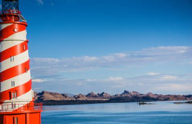 Büyük bir su kütlesine tepeden bakan kayalık bir kıyı şeridinde kırmızı ve beyaz çizgili bir deniz feneri var. Gökyüzü açık ve mavi, ve uzaktaki dağlar dingin atmosfere katkı yapıyor.