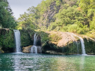 Arka planda yemyeşil bir orman olan bir şelale nehre akıyor. Su berrak ve sakin, manzara huzurlu ve huzurlu.