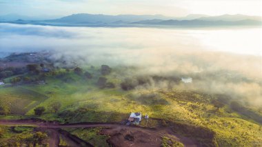 Sisli, sisli bir sabah ve tarlanın ortasında küçük bir ev. Gökyüzü bulutlu ve manzara huzurlu.