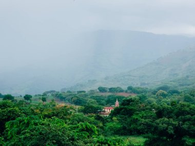 Puslu dağlar yemyeşil vadilerin üzerinde beliriyor. Küçük kırmızı çatılı ev ağaçların arasına kuruludur. Sis, tepeleri usulca örterek manzarayı yumuşatıyor. Titreyen yapraklar sisli, sakin bir gökyüzünün altında sallanıyor.