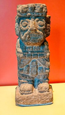 Antik taş heykel ciddiyetle kaidede duruyor. Oyulmuş yüz, karmaşık gözler ve kaşlar gösteriyor. Mavi pigmentler aşınmış, desenli yüzey. Kültürel eser gizem ve tarih yayar.