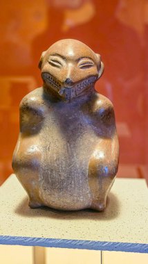 Eski çömlek figürü sakince sergilenir. Yumuşak kıvrımlar onun neşeli, gülen yüzünü tanımlar. Sönük tonlar, sıcak arka plan ışıklandırmasıyla tezat oluşturuyor. Kıvrımlı eller nazik, misafirperver duruşu gösterir.