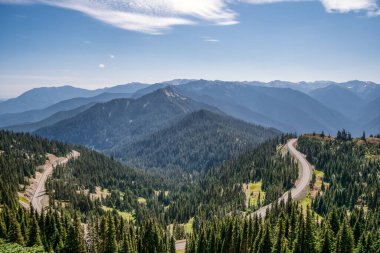 Dağ yolu sık ormanda esiyor. Gökyüzü açık mavi, bulutlar yavaşça süzülüyor. Evergreen ağaçları yuvarlanan yamaçlar boyunca uzanıyor. Yol doğanın kucağında zarifçe kıvrılır.