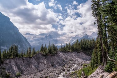 Engebeli dağların üzerinde dramatik bulutlar uçuşuyor. Karla kaplı tepeler hep yeşil ağaçların arkasında dikilir. Rocky yamaçları sığ bir akıntıya doğru kayıyor. Ormanın derinliklerindeki gölgeler vadinin vahşi güzelliğini çerçeveler.