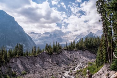 Karla kaplı zirveler engebeli bir uçurumun arkasında beliriyor. Kozalaklı ağaçlar dik, kayalık inişi çerçeveler. Dağınık bulutlu puslu gökyüzü derinlik katıyor. Doğanın ham gücü dramatik manzaraya hükmediyor.