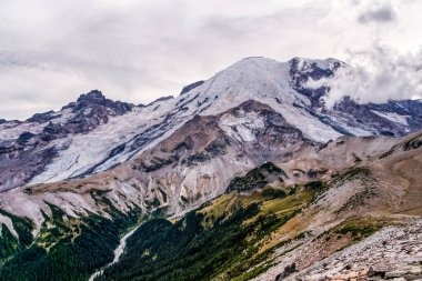 Karla kaplı tepe engebeli kayalık arazide yükselir. Yeşil vadiler dik, güneşli yamaçlardan esiyor. Bulutlar yavaşça zirvenin sivri kenarına doğru sürükleniyor.