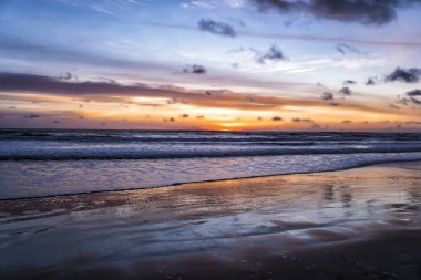 Güneş alçalıyor, gökyüzünü turuncu ve mor renklere boyuyor. Dalgalar hafifçe çarpar, ıslak kumda parlar. Bulutlar yavaşça sürükleniyor, altın ufku çerçeveliyor. Sessiz sahil sakin, davetkar dinginlik