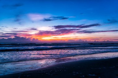 Derin mavi gökyüzü okyanus ufkunda turuncuya döner. Yumuşak dalgalar, sessiz ritimle siyah kumu kaplar. Bulutlar pembe ve mor parlıyor, ıslak kumsala yansıyor. Günbatımı gökyüzünü zengin, katmanlı renklerle boyar.