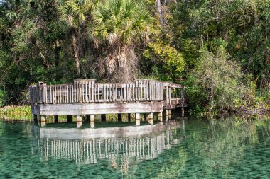 Ahşap iskele sakin ve berrak suyun üzerine uzanır. Yansımalar yüzeyin altında parıldıyor. Olay yerini avuçlar ve yeşillikler sarmış durumda. Huzurlu atmosfer sessiz tefekküre davet eder.